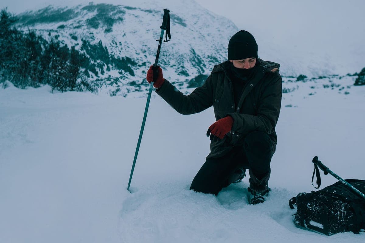 A person kneeling in a snowy mountain setting holding a trekking pole