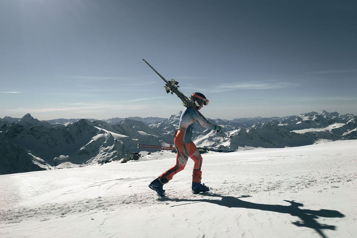 A skier carrying skis over one shoulder on a snowy mountain with alpine peaks behind