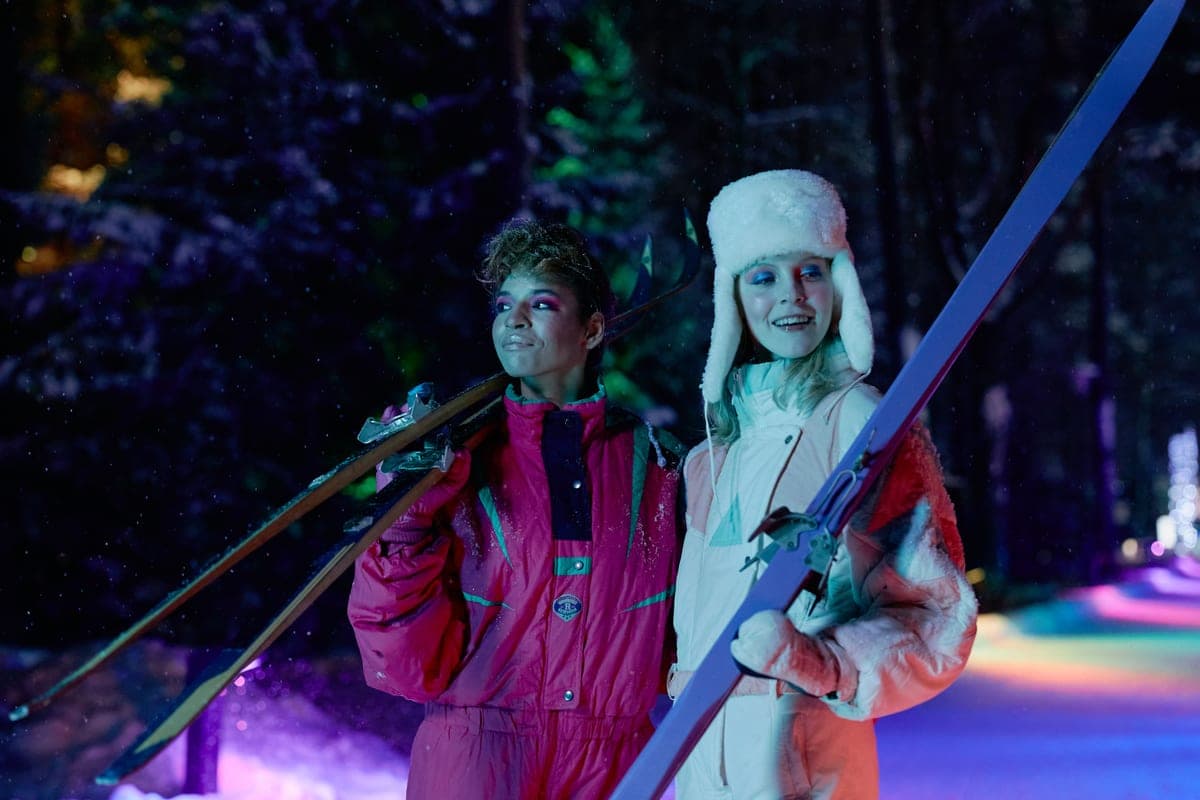 Two people in winter clothing holding skis in the snow at night