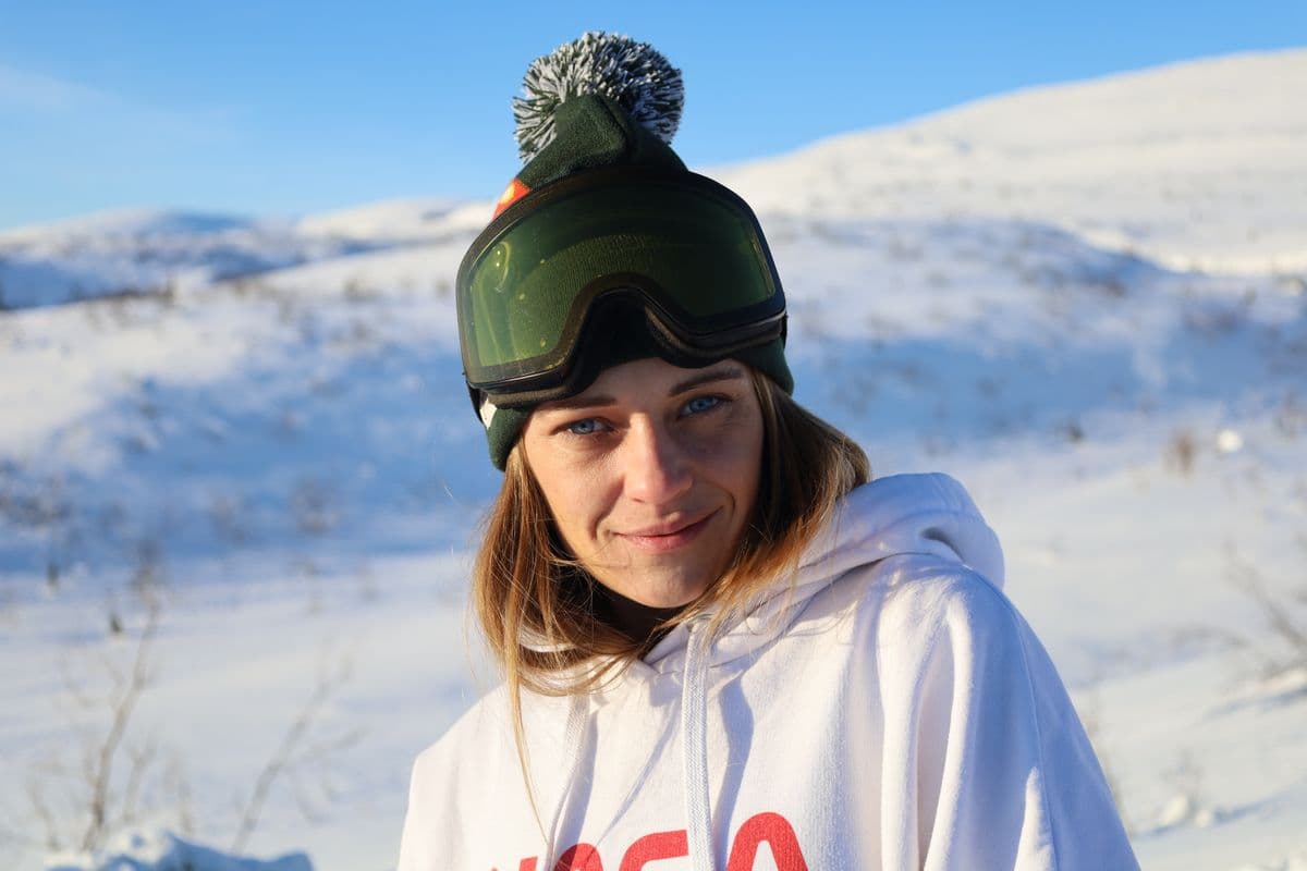 A woman wearing goggles and a hood smiling against a snowy mountain backdrop